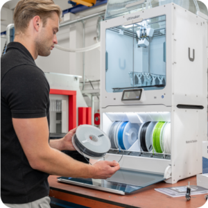 Man loading filament into an Ultimaker 3D printer in a modern workshop, with spools of material and Ultimaker packaging visible on the workbench.